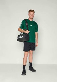 Πράσινη μπλούζα Adidas με μαύρες λεπτομέρειες, μαύρες βερμούδες και μαύρη αθλητική τσάντα. Κοντά μανίκια και άνετη εφαρμογή. Μαύρα αθλητικά παπούτσια.