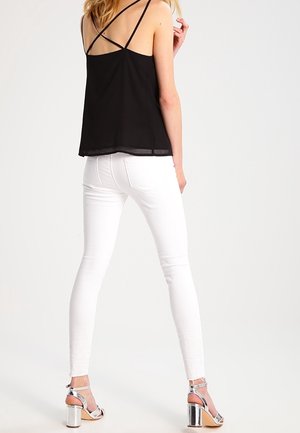 Top nero senza maniche con spalline incrociate, abbinato a jeans bianchi aderenti e sandali con tacco argento, che mostrano texture lisce e linee pulite.