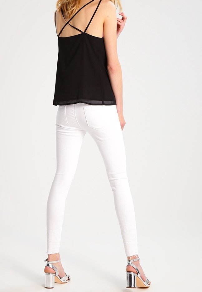 Top nero senza maniche con spalline incrociate, abbinato a jeans bianchi aderenti e sandali con tacco argento, che mostrano texture lisce e linee pulite.