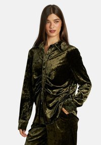 Camisa de terciopelo verde oscuro con mangas largas, cierre frontal de botones y una textura suave y lisa. Cuenta con un collar clásico y detalles fruncidos.