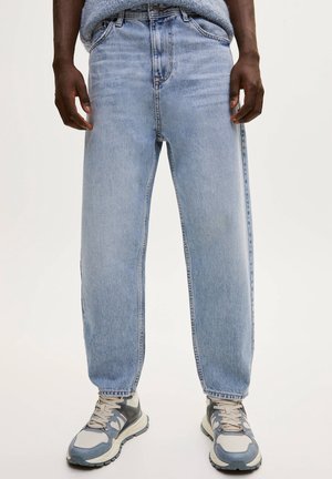 Osoba ubrana w jasnoniebieskie, luźne dżinsy i szare trampki, stojąca na tle jednolitego tła.