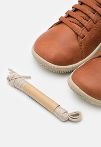 Braune Ledersneakers mit strukturiertem Finish, ausgestattet mit beige Schnürsenkeln und einer Gummisohle. Ein aufgerollter beiger Schnürsenkel liegt neben den Schuhen.