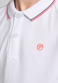 Valkoinen polo paita, jossa on teksturoitu kangas, vaaleanpunainen ja musta raidallinen kaulus, kaksi nappia ja pieni vaaleanpunainen logo rinnassa.