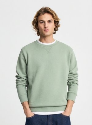 Giovane uomo con capelli biondi mossi che indossa una felpa verde chiaro e jeans blu, in piedi con le mani nelle tasche davanti a uno sfondo semplice.