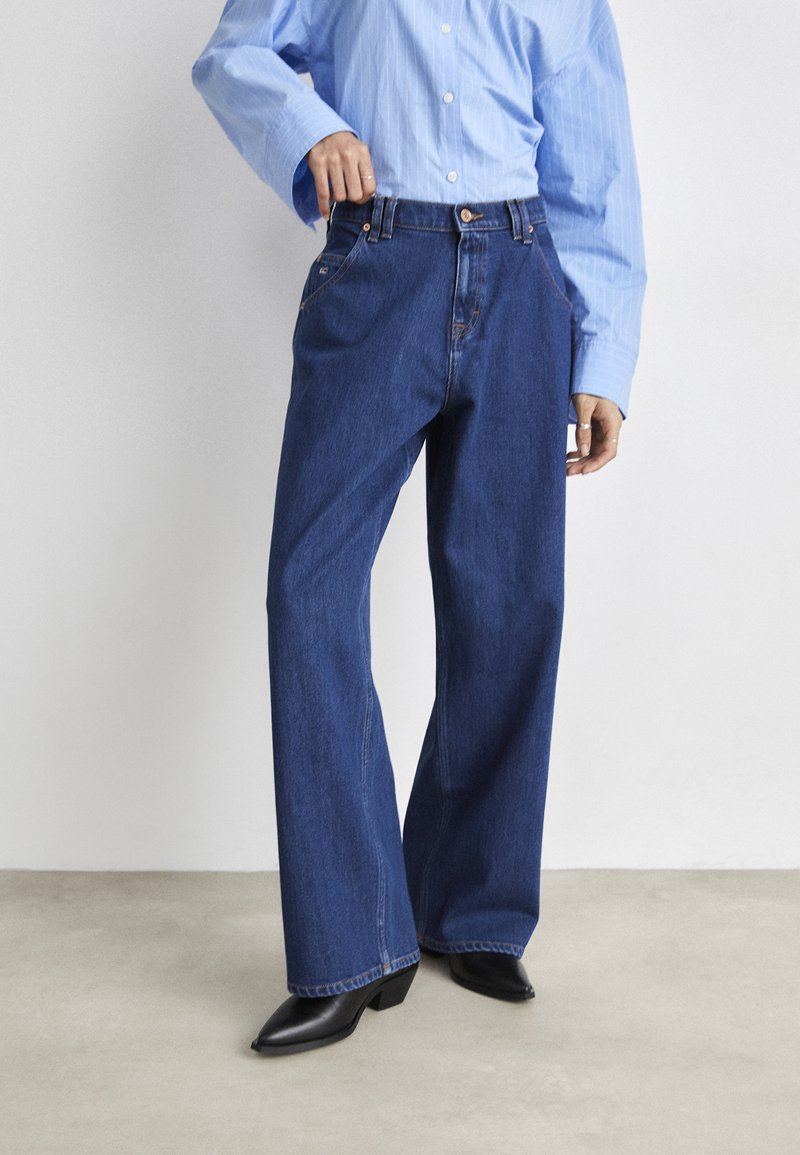 Bredde ben bukser i blå denim med høy midje, med fem lommer og klassisk knappestolpe. Kombinert med en lyseblå skjorte med knapper.