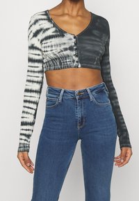 Gekürztes langärmliges Oberteil in Schwarz-Weiß-Tie-Dye, mit tiefem V-Ausschnitt und Knopfleiste. Kombiniert mit hochgeschnittenen blauen Jeans.