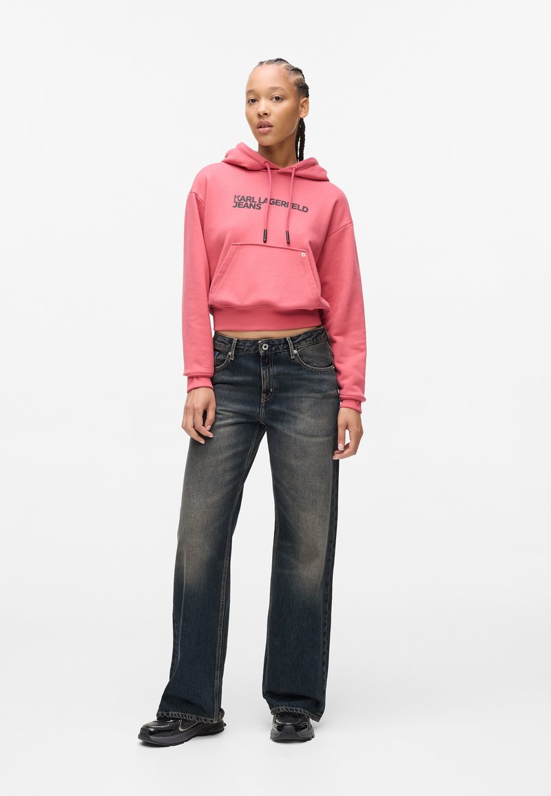Rosa kurz geschnittenes Hoodie mit Fronttasche und schwarzem Logo, kombiniert mit dunkelblauen weit geschnittenen Jeans mit ausgewaschenem Finish und Gürtelschlaufen.