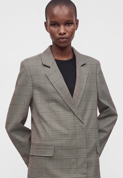 Personne portant une chemise noire et un blazer à carreaux surdimensionné, les mains dans les poches, debout devant un fond clair uni.