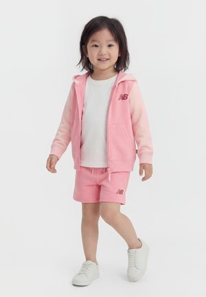 Niño pequeño de pie, vestido con sudadera rosa con cremallera, pantalones cortos a juego, camiseta blanca y zapatillas blancas, sonriendo a la cámara.