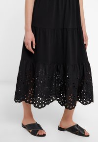 Černé vrstvené šaty s otevřeným květinovým vzorem na spodním lemu, doplněné černými klínovými sandály se špičatými ozdobami a křížovými řemínky.