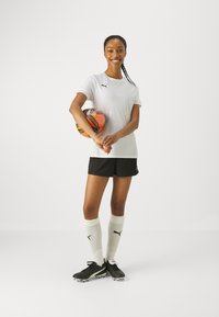 Hvit sporty skjorte med svart logo, svarte shorts, svarte fotballsko, og hvite knelange sokker. Holder en oransje og blå fotball.
