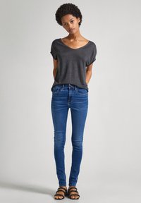 Camiseta gris de cuello en V combinada con jeans ajustados azules. Los jeans tienen un diseño clásico de cinco bolsillos. Se lleva con sandalias negras de tiras.