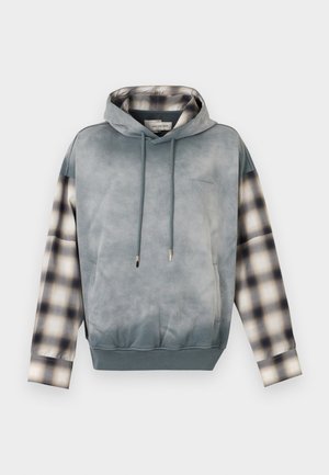 Fiatal férfi szürke pulóverben kockás ujjakkal és barna, bő szárú nadrágban, amelynek oldalsó panelein kockás minta található, zsebre tett kézzel áll.