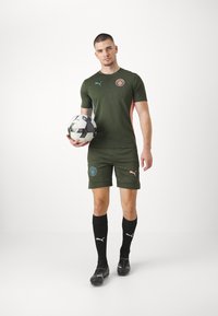 Tumšu zaļu Puma sporta komplektu, kas ietver teksturētu augšu ar rozā akcentiem, saskaņotas šortus, melnas zeķes un futbolu, uz baltas fonu.
