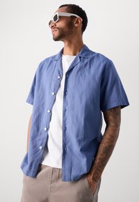 Camicia azzurra a maniche corte con bottoni frontali, abbinata a una t-shirt bianca. Presenta una vestibilità rilassata e una texture di tessuto morbida.