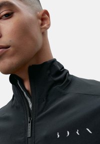 Sort zip-up jakke lavet af glat stof; har en høj krave og kontrasterende hvid logo detalje nær lynlåsen.