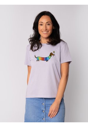 Frau mit dunklem Haar, die lächelt, trägt ein hellviolettes T-Shirt mit einem bunten, gestreiften Dackel, der eine gelbe Blume hält, und einen blauen Rock mit Knöpfen.