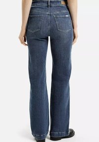 Jeans z wysokim stanem w ciemnoniebieskim deninie, z dwoma tylnymi kieszeniami i teksturowanym materiałem o lekkim połysku, noszone z czarnymi butami.