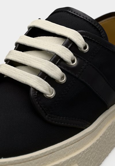 Κοντινό πλάνο μαύρου αθλητικού παπουτσιού με λευκά, παχιά κορδόνια που περνούν μέσα από μεταλλικά ρημάδια και με υφασμένο, εκρού πέλμα.