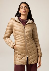 Beige pufferjack met chevronstiksel, ritssluiting en een capuchon. Lichtgewicht, glanzende stof met geribbelde textuur en zijzakken.