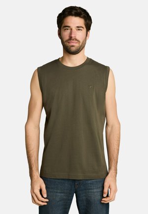 Man draagt een mouwloos olijfgroen shirt en blauwe spijkerbroek, staand tegen een effen lichte achtergrond.