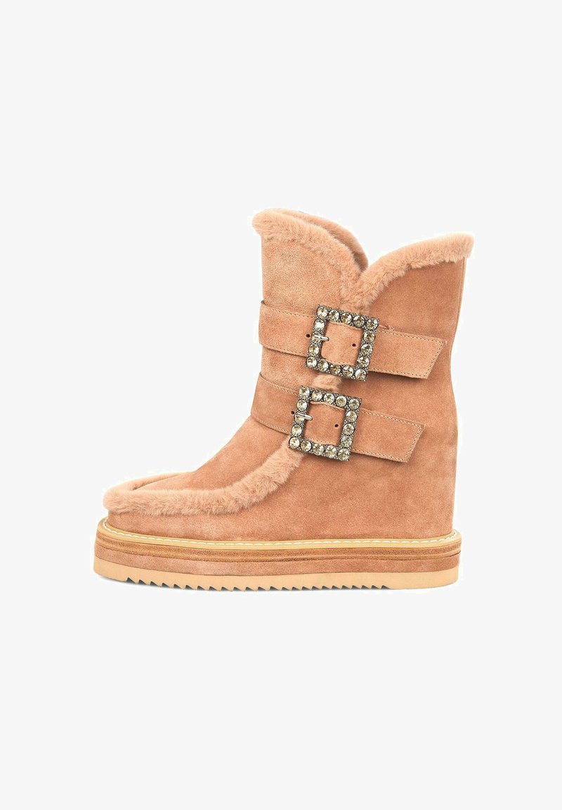 Botas de ante marrón con ribete de piel de oveja, dos hebillas adornadas con cristales y suela de cuña. Presenta una puntera redondeada y suela texturizada.