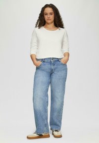 Top bianco a maniche lunghe leggero, vestibilità comoda. Jeans blu a gamba larga con finitura sbiadita, tasche frontali, indossati con sneakers marroni e beige.