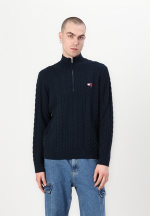 Maglione lavorato a maglia blu navy con colletto alto, zip e motivi testurizzati. Presenta una piccola toppa con logo sul lato sinistro del petto. Vestibilità casual.