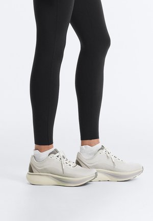 Osoba ubrana w białe sportowe trampki i czarne legginsy stojąca na białej powierzchni na tle jasnoszarym.