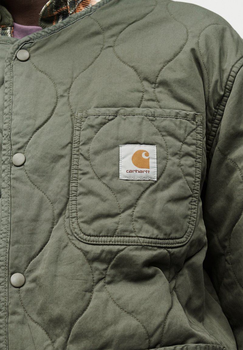 Veste matelassée verte avec poche avant ornée d'un patch logo Carhartt et fermeture à boutons pression.