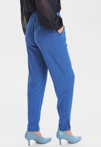 Kobieta ubrana w wzorzyste niebieskie spodnie z bocznymi kieszeniami i dekoltem w szwie, zestawione z jasnoniebieskimi szpilkami oraz przezroczystą czarną bluzką w niebieskie kropki.