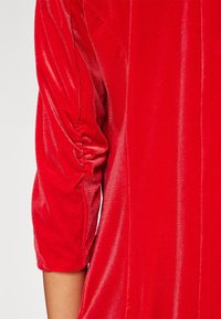 Vêtement en velours rouge à manches trois-quarts. Le tissu a une texture douce et brillante avec des détails froncés sur les manches.