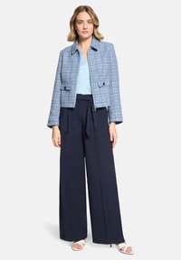 Femme portant une veste en tweed bleu clair, un chemisier bleu pâle, un pantalon large bleu marine avec ceinture, et des talons ouverts blancs, debout devant un fond blanc.
