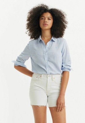 Chemise à rayures bleu clair, manches retroussées, associée à un short en denim blanc taille haute avec un ourlet effiloché.
