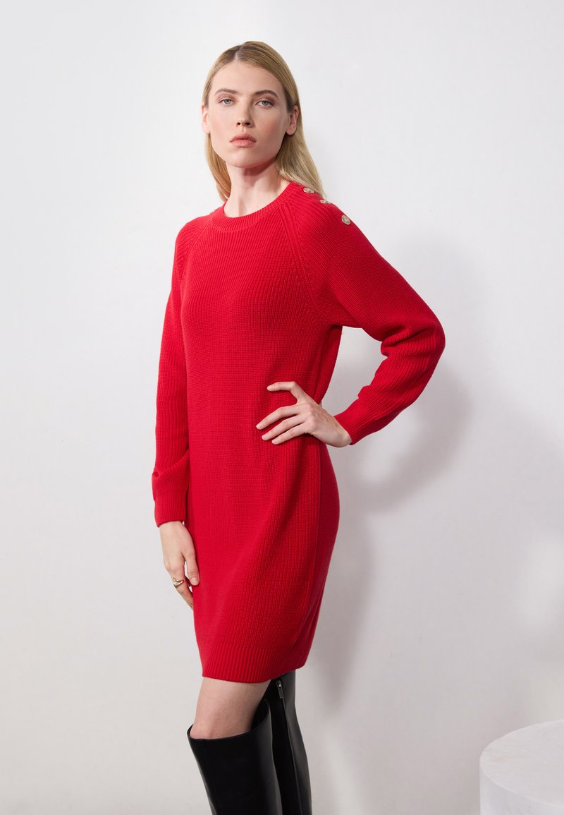 Robe tricotée rouge à manches longues, texture côtelée et accents de boutons sur l'épaule. Elle a une coupe décontractée, atteignant la mi-cuisse.