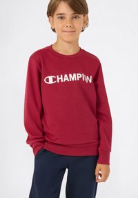 Sudadera roja hecha de mezcla de algodón, con mangas largas y cuello redondo. Logo blanco de "CHAMPION" exhibido de manera prominente en el frente.