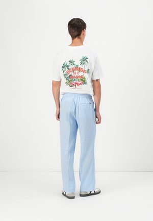 Homme vu de dos portant un T-shirt blanc avec un imprimé voiture tropicale et un pantalon bleu clair sur fond blanc uni.