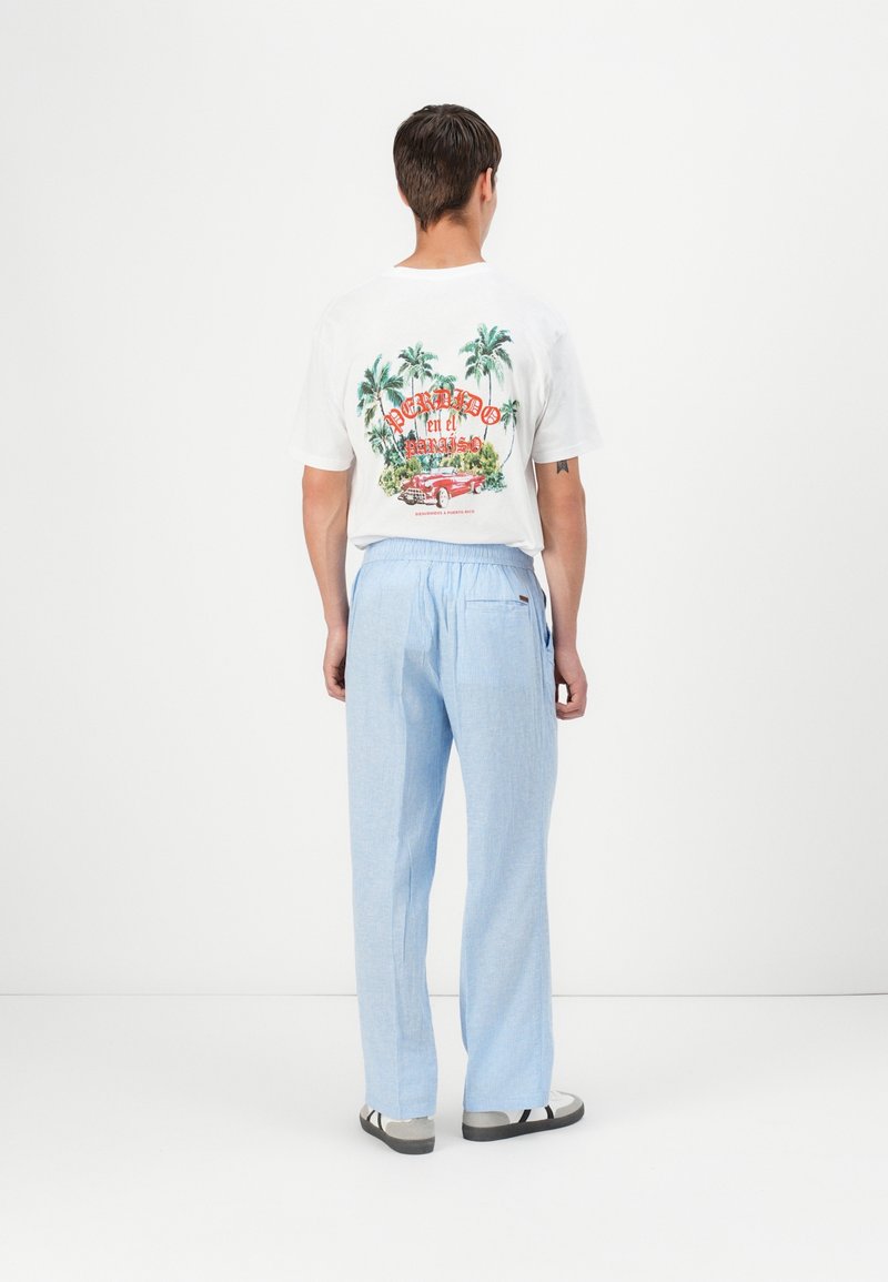 Homme vu de dos portant un T-shirt blanc avec un imprimé voiture tropicale et un pantalon bleu clair sur fond blanc uni.