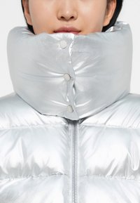 Silver puffer jacket with a high, padded collar, featuring snap buttons and a central zipper. Smooth, reflective texture.
