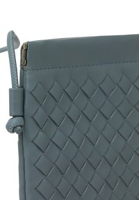 Grå läderclutch med vävt mönster, slät textur och dragkedjestängning. Har en liten dekorativ ögla på sidan.