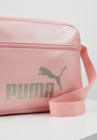 Borsa a tracolla in ecopelle rosa con logo Puma argentato, chiusura con zip e tracolla in tessuto morbido. Texture liscia e forma rettangolare.