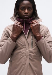 Άτομο που φοράει μια μπεζ αδιάβροχη ζακέτα με κουκούλα πάνω από μια μπορντό ζακέτα με φερμουάρ, κρατώντας το γιακά της ζακέτας με τα δύο χέρια.