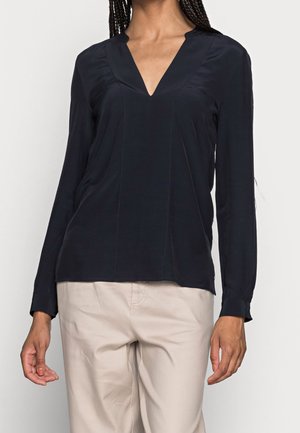 Femme portant un chemisier noir à manches longues et col en V ainsi qu’un pantalon beige clair, debout, les bras détendus le long du corps, sur un fond clair uni.