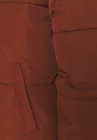 Veste matelassée marron avec un extérieur lisse et mat. Comprend une poche latérale proéminente et des coutures discrètes le long des coutures.