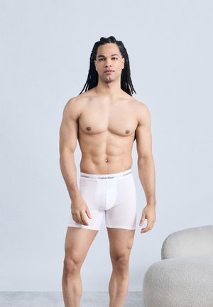 Μυώδης άνδρας με μακριά πλεγμένη τρίχα στέκεται φορώντας λευκά μπόξερ Calvin Klein μπροστά σε απλό ανοιχτόχρωμο φόντο.