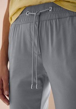 Close-up van een persoon die grijze broek met trekkoord en elastische taille en een geel shirt draagt, hand rust langs het lichaam.