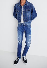 Džínová bunda se světle modrým praním, dvě přední kapsy a kovové knoflíky, v kombinaci s opranými modrými džíny a černými teniskami.