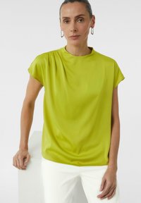 Bluzka w kolorze limonki z wysokim kołnierzem, krótkimi rękawami w formie kaptura i zwiewnym krojem, zestawiona z białymi, dopasowanymi spodniami. Gładka tekstura tkaniny.