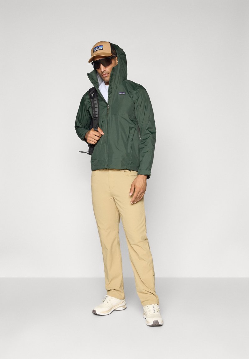 Homme portant une veste à capuche verte, un pantalon beige, des baskets blanches, des lunettes de soleil et une casquette marron, se tenant avec un sac à dos noir sur une épaule.