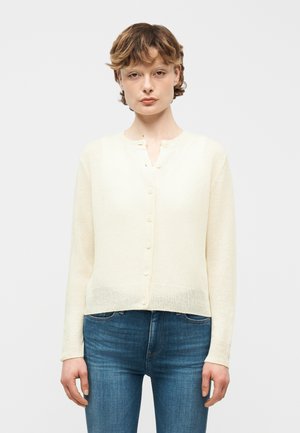 Young person with short wavy hair wearing a cream buttoned cardigan and blue jeans, standing against a plain light background.